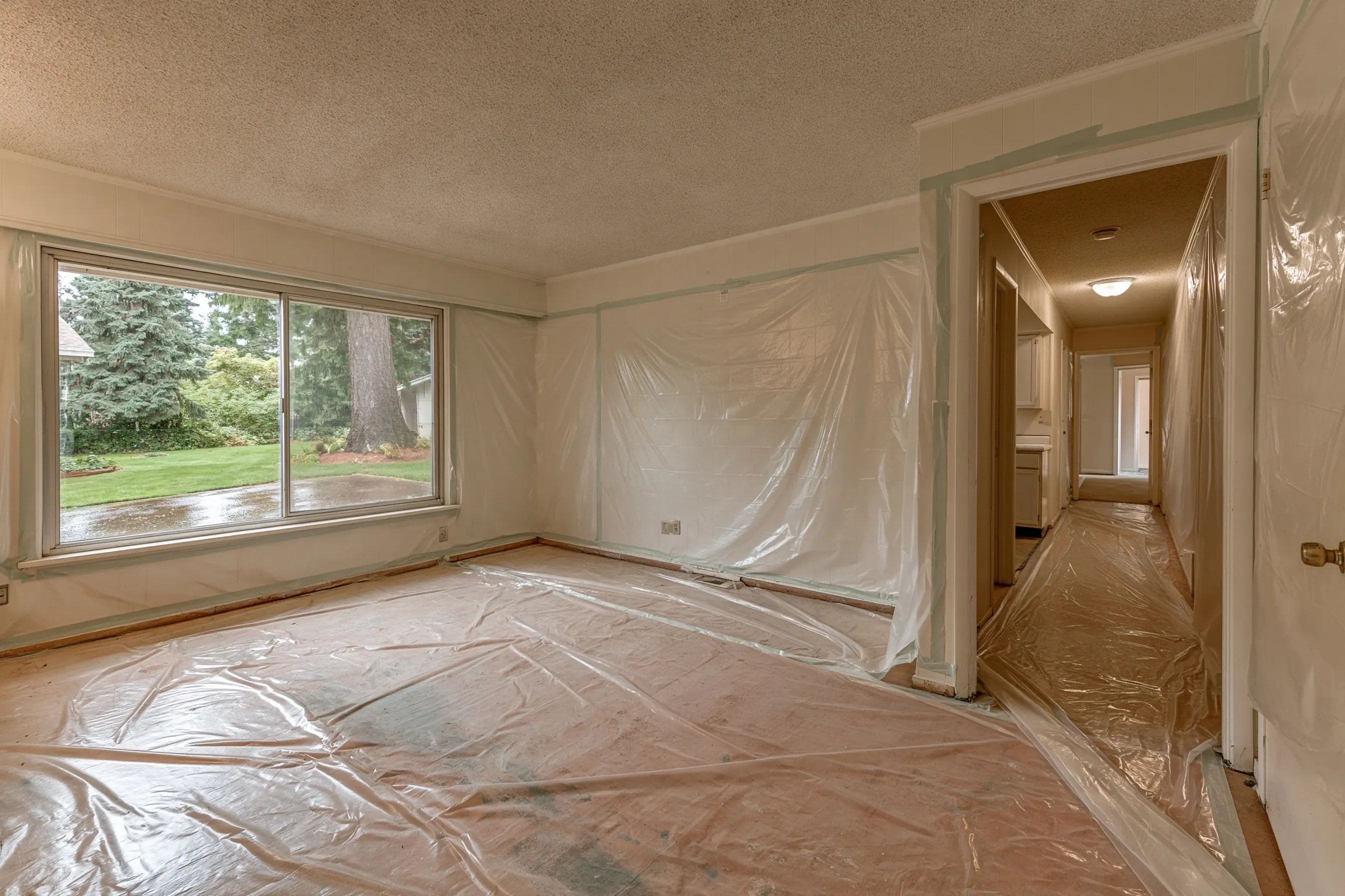 Rambler living room wrapped in plastic under a bumpy ceiling, with a wide slider window to a rainy lawn and a sealed hallway beyond